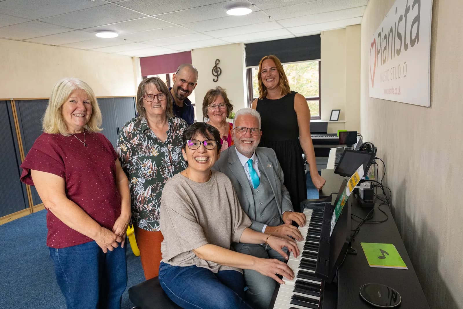 A group of seven adults, including two playing a digital piano, pose and smile in a music studio.