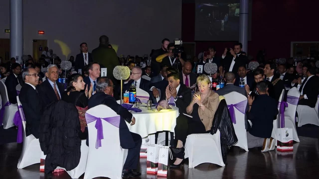 A large group of people in formal attire are seated around tables with white tablecloths and purple bows on chairs, attending an event in a dimly lit hall, with some individuals looking towards the camera.